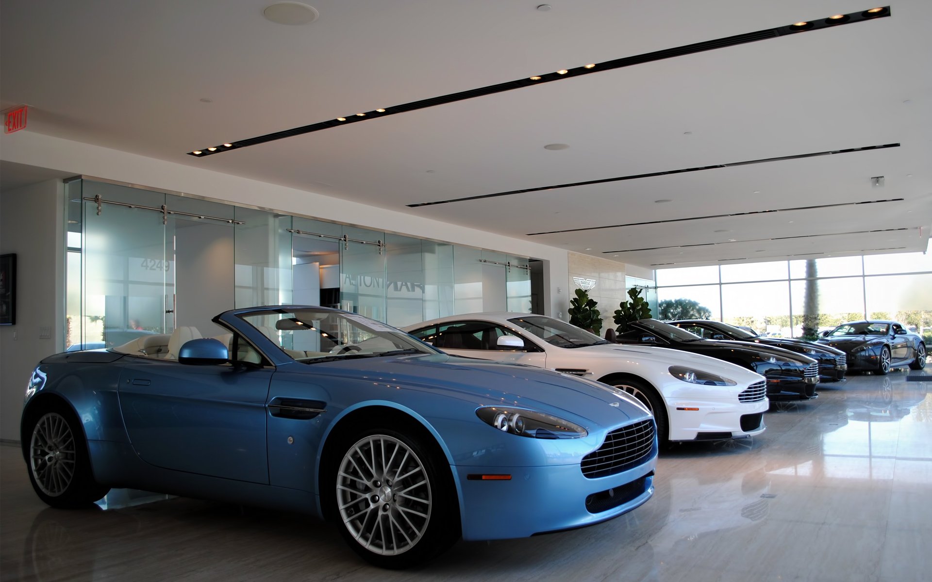 HD PC desktop wallpaper showing a row of Aston Martin sports cars in a bright showroom, foreground sky-blue convertible.