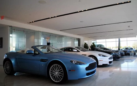HD PC desktop wallpaper showing a row of Aston Martin sports cars in a bright showroom, foreground sky-blue convertible.