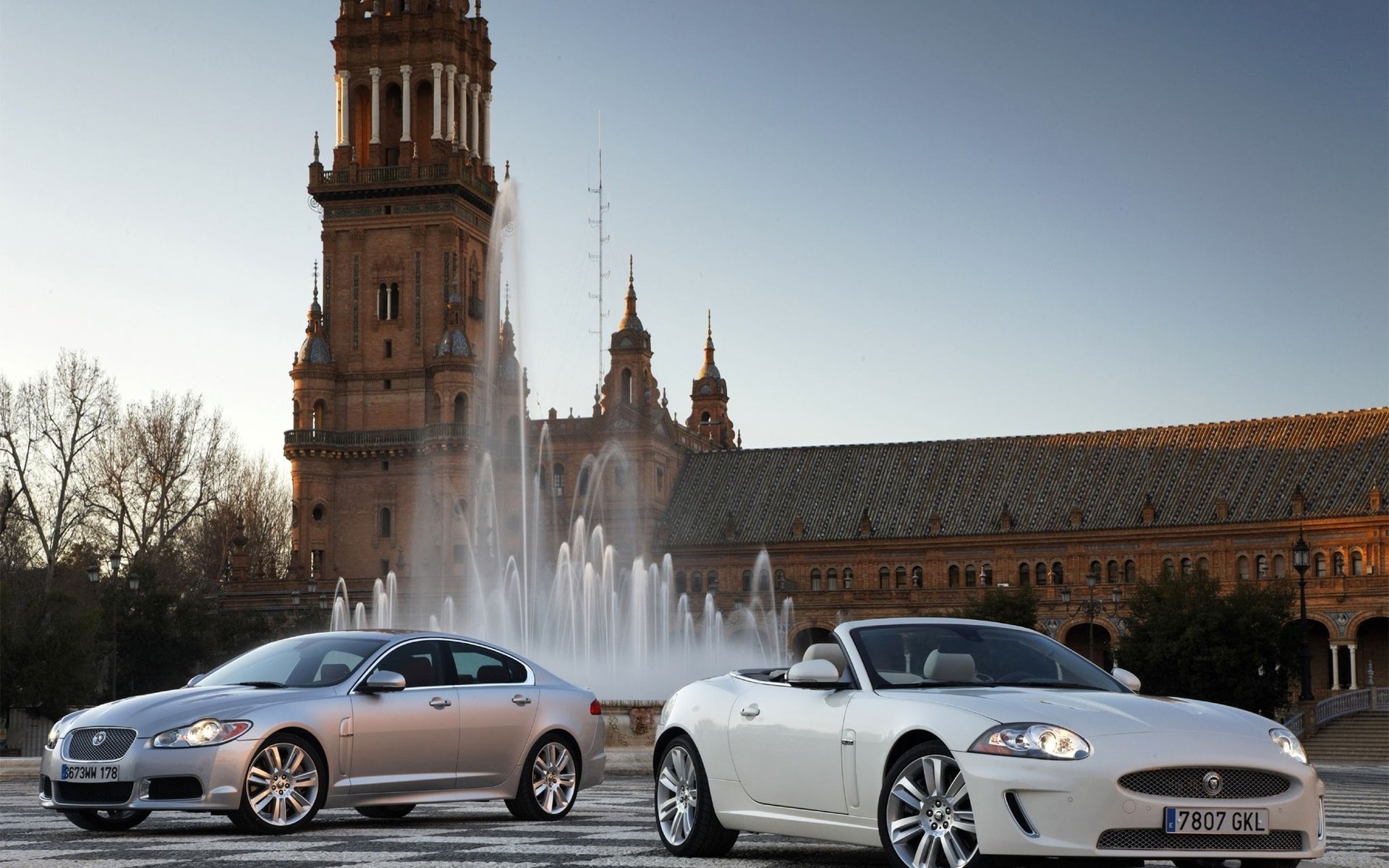 HD PC desktop wallpaper: two silver Jaguar cars parked before a fountain and ornate plaza tower at dusk.