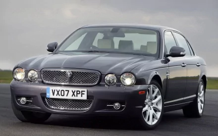 HD PC desktop wallpaper of a grey Jaguar vehicle sedan, three-quarter front view with chrome grille and alloy wheels on an open tarmac background.