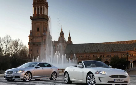 HD PC desktop wallpaper: two silver Jaguar cars parked before a fountain and ornate plaza tower at dusk.
