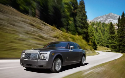 HD PC desktop wallpaper/background: a silver Rolls-Royce coupe driving on a winding mountain road past pine forests under a clear blue sky.