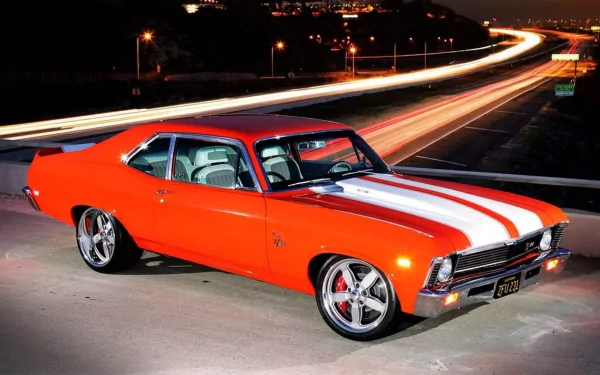 HD PC desktop wallpaper showing a classic Chevrolet muscle car in bright orange with white racing stripes, parked against a night backdrop with light trails on a highway.