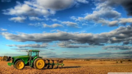 HD desktop wallpaper featuring a green John Deere tractor working on a vast, sunlit field under a blue sky with scattered clouds.