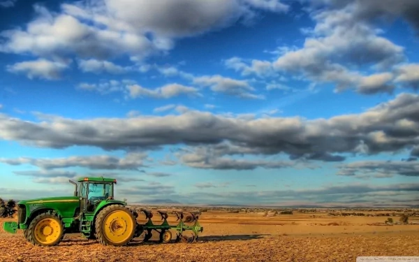 HD desktop wallpaper featuring a green John Deere tractor working on a vast, sunlit field under a blue sky with scattered clouds.