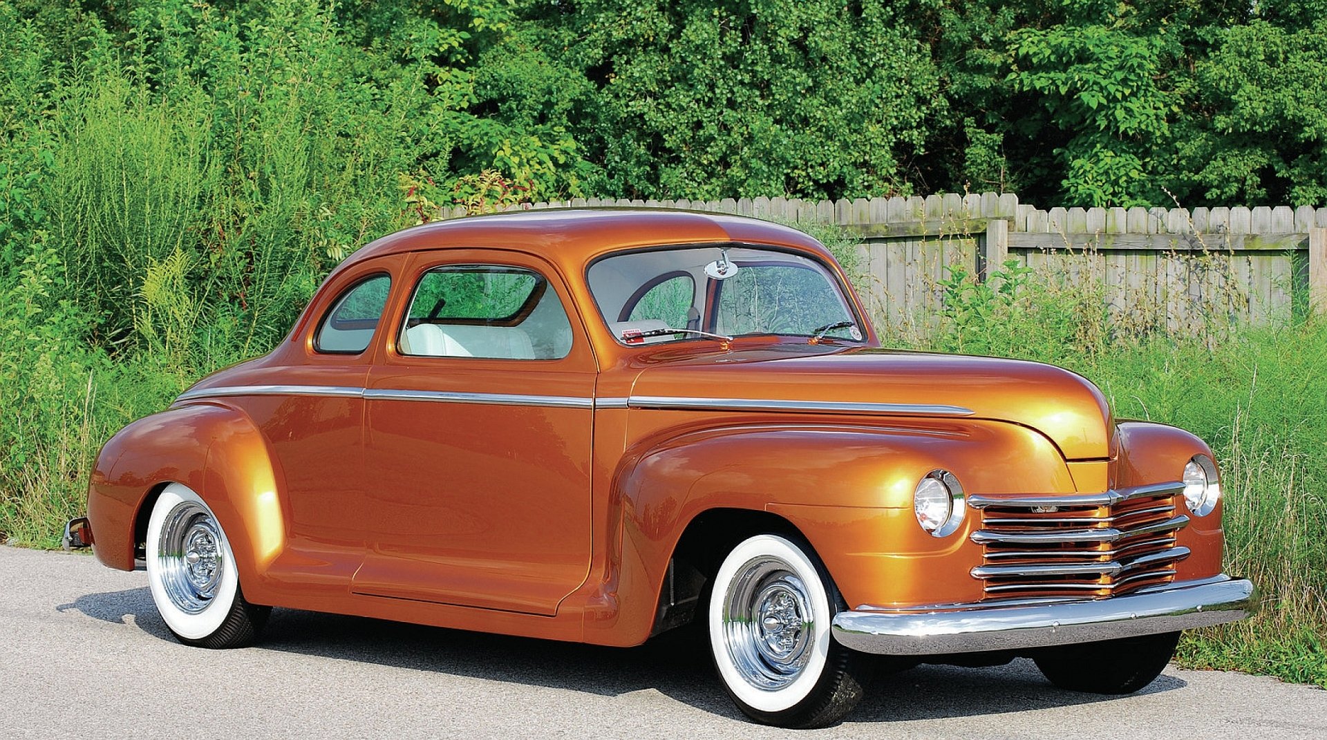 HD desktop wallpaper featuring a classic orange Plymouth vehicle parked outdoors against a backdrop of greenery and a wooden fence.