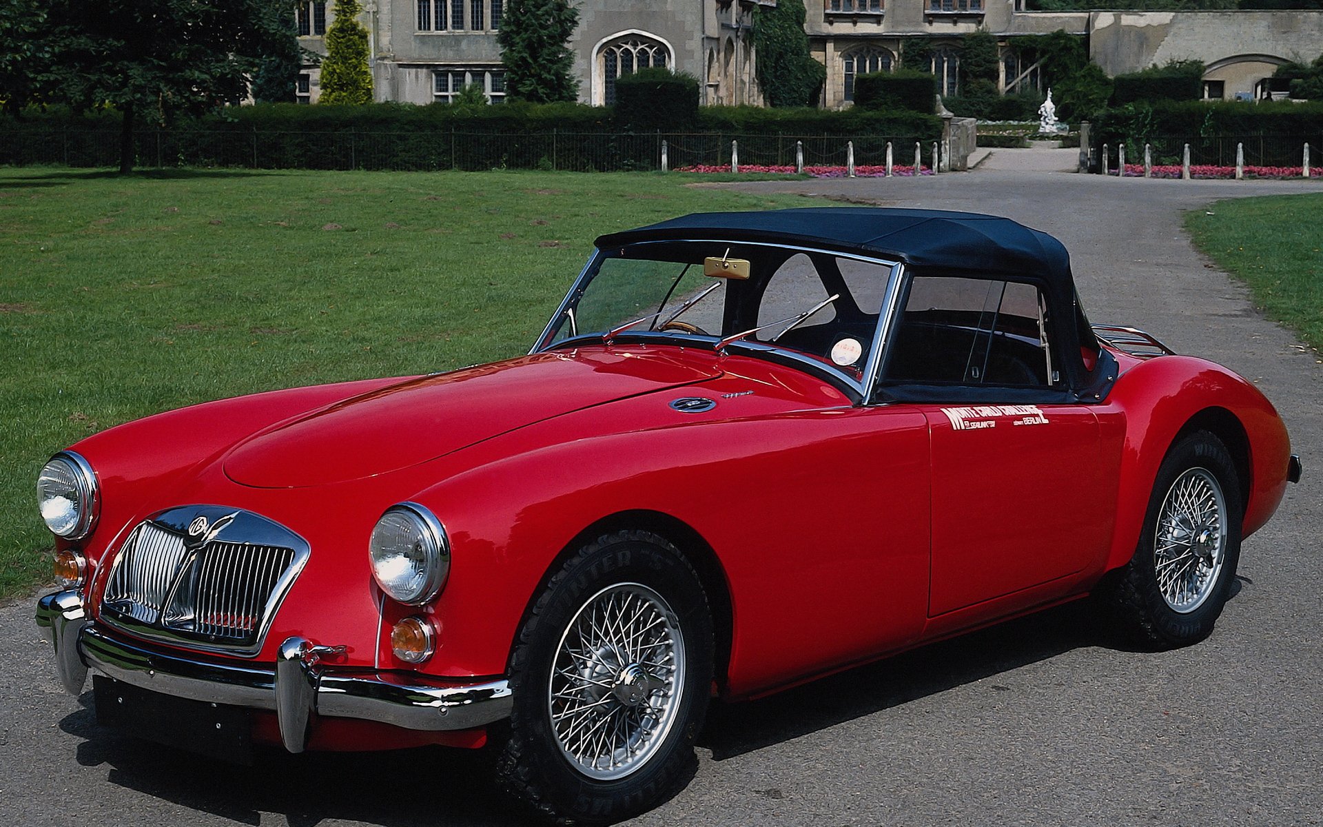 HD PC desktop wallpaper featuring a classic red MG convertible vehicle parked on a driveway with a large building and green lawn in the background.