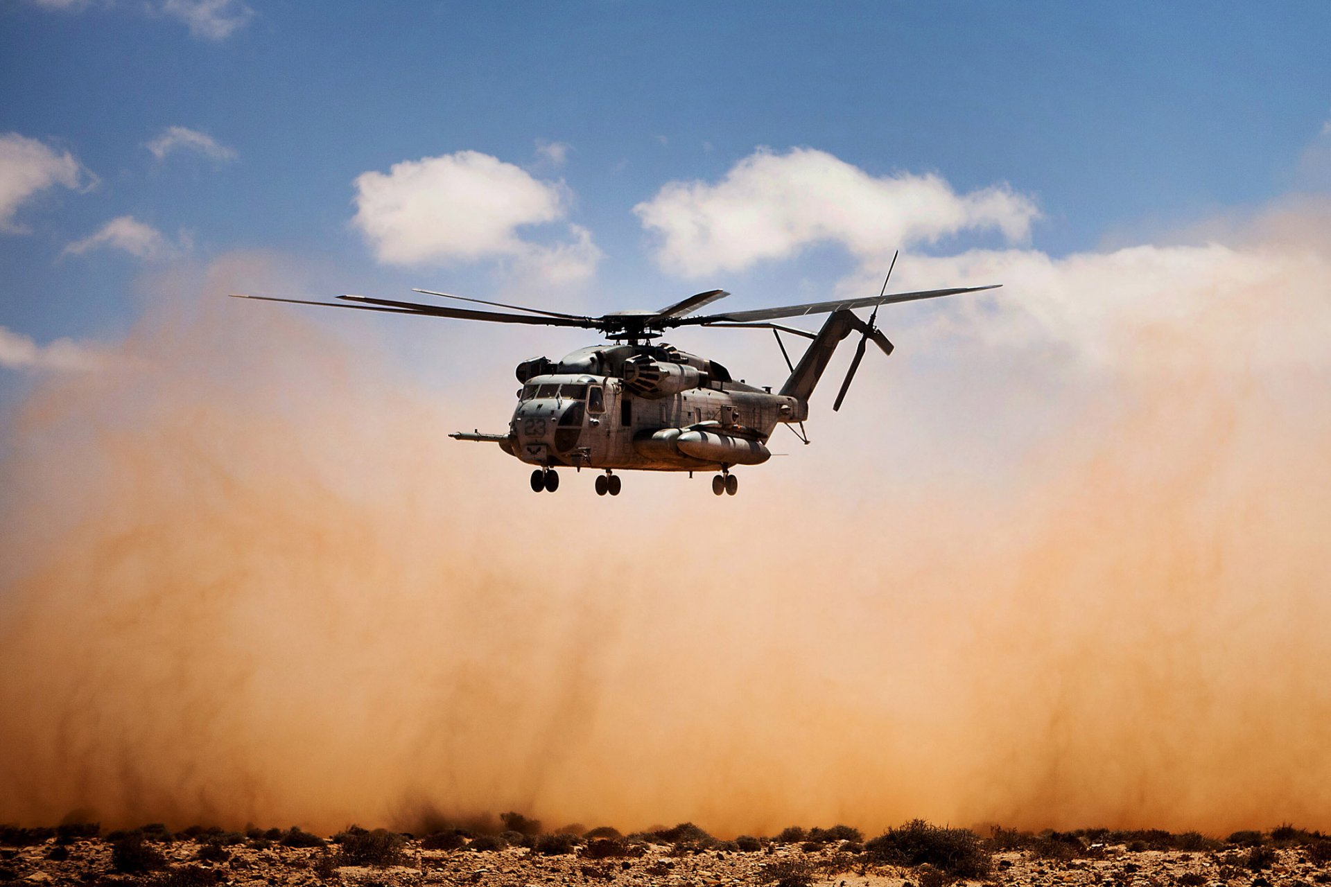 HD military desktop wallpaper featuring a Sikorsky CH-53E Super Stallion helicopter hovering above dusty terrain under a blue sky with scattered clouds.