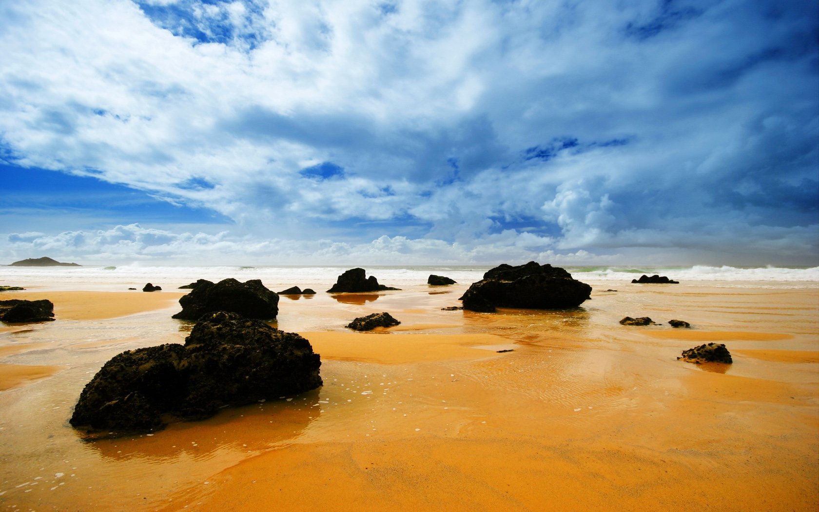 Vibrant HD desktop wallpaper of a serene beach landscape, showcasing golden sand, scattered rocks, a vast ocean, and a dramatic sky filled with clouds.