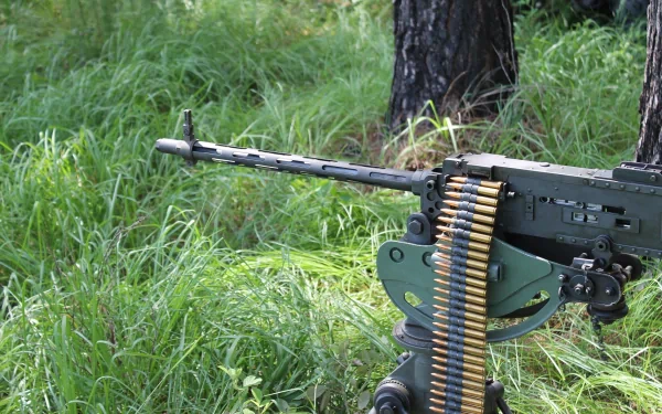 A close-up view of a man-made machine gun positioned on a stand, surrounded by lush green grass, creating a striking HD desktop wallpaper and background.