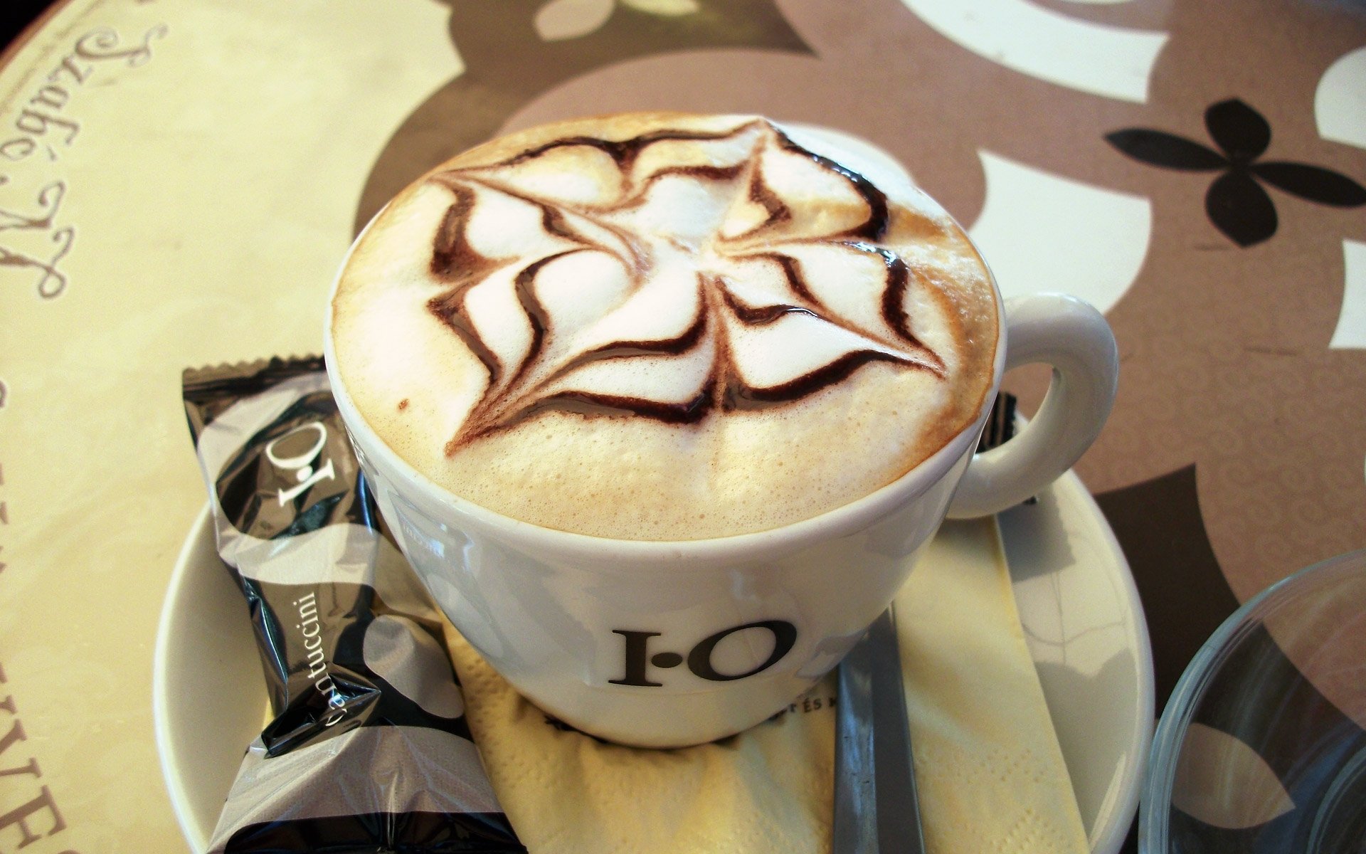 HD PC desktop wallpaper showing coffee: a cappuccino with chocolate star latte art on a saucer with a cookie and wrapped biscuit on a decorative tabletop.