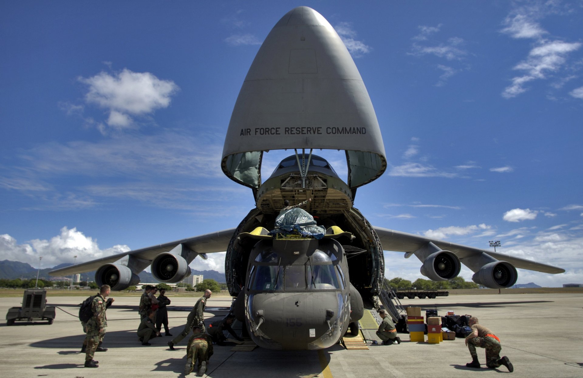 Lockheed C-5 Galaxy with open nose cargo bay loading a Boeing CH-47 Chinook helicopter, surrounded by Air Force personnel on a sunny military airbase.