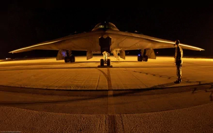 A stealth aircraft sits on the runway at night, illuminated by soft lights, with a military personnel standing nearby, highlighting its sleek design and advanced technology.
