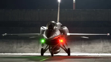 A General Dynamics F-16 Fighting Falcon stands on the tarmac at night, illuminated by vibrant green and red lights, showcasing its sleek design and military prowess.