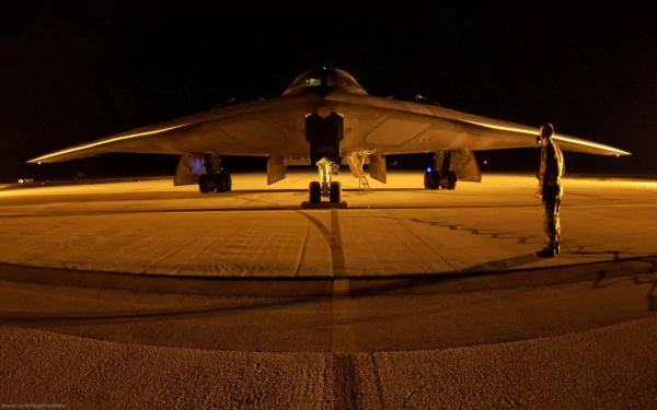 A stealth aircraft sits on the runway at night, illuminated by soft lights, with a military personnel standing nearby, highlighting its sleek design and advanced technology.