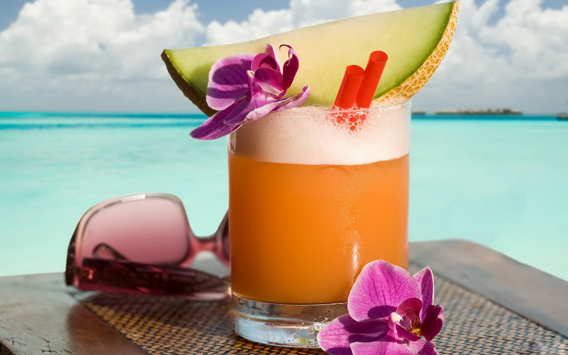 HD PC desktop wallpaper of a tropical orange cocktail garnished with a lime wedge and orchid, set against a turquoise ocean background with pink sunglasses nearby.
