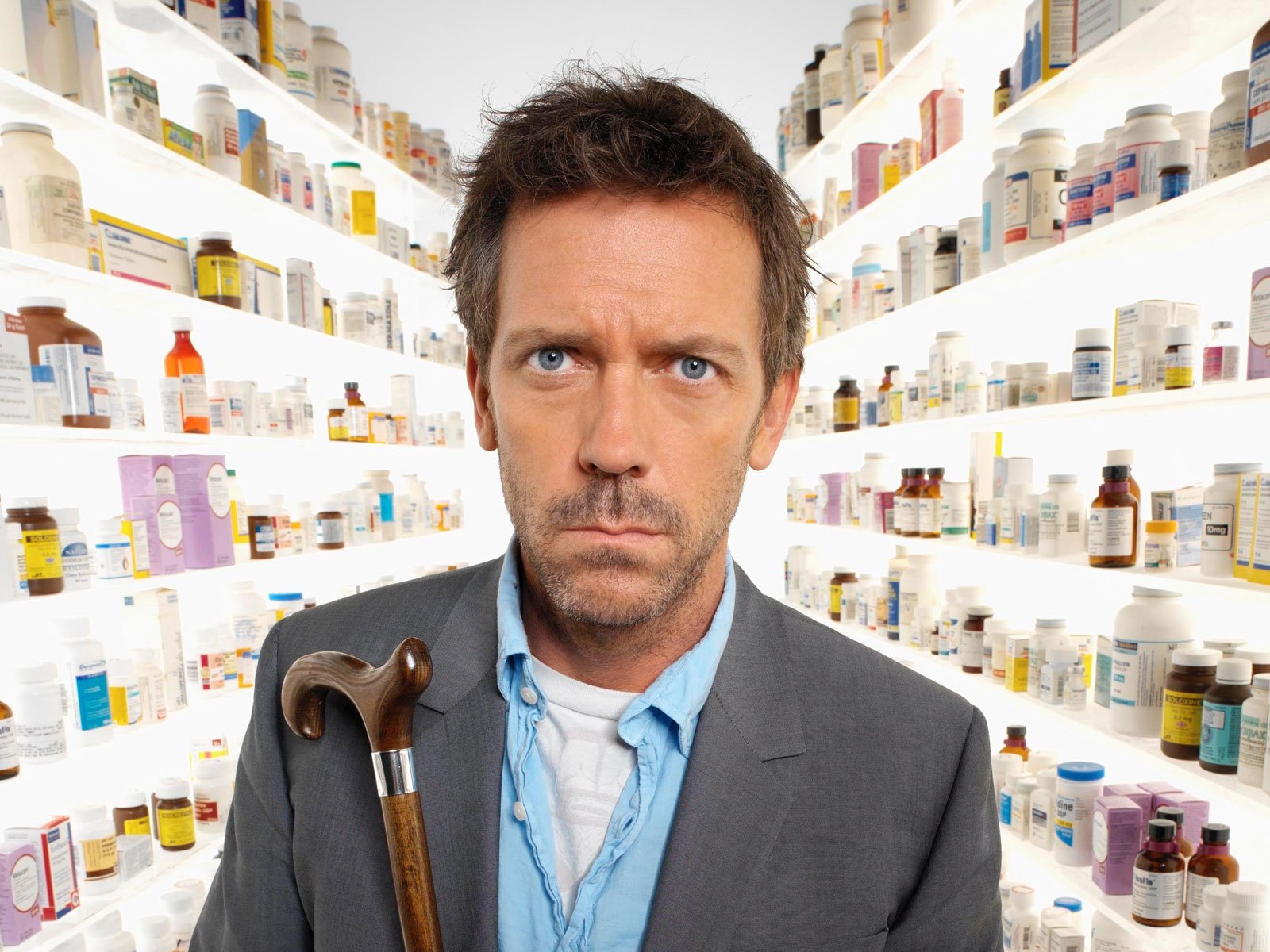 HD desktop wallpaper featuring Gregory House, portrayed by Hugh Laurie, from the TV show House, standing in front of shelves filled with medical supplies, holding a cane.
