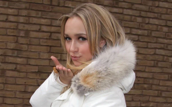Hayden Panettiere blowing a kiss, dressed in a stylish white coat with a fur hood, against a textured brick background in this striking HD desktop wallpaper.