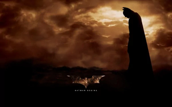 HD desktop wallpaper of a Batman Begins scene, featuring Batman silhouetted against a dramatic, cloudy sky. The background highlights the iconic Batman symbol and the movie title at the bottom.
