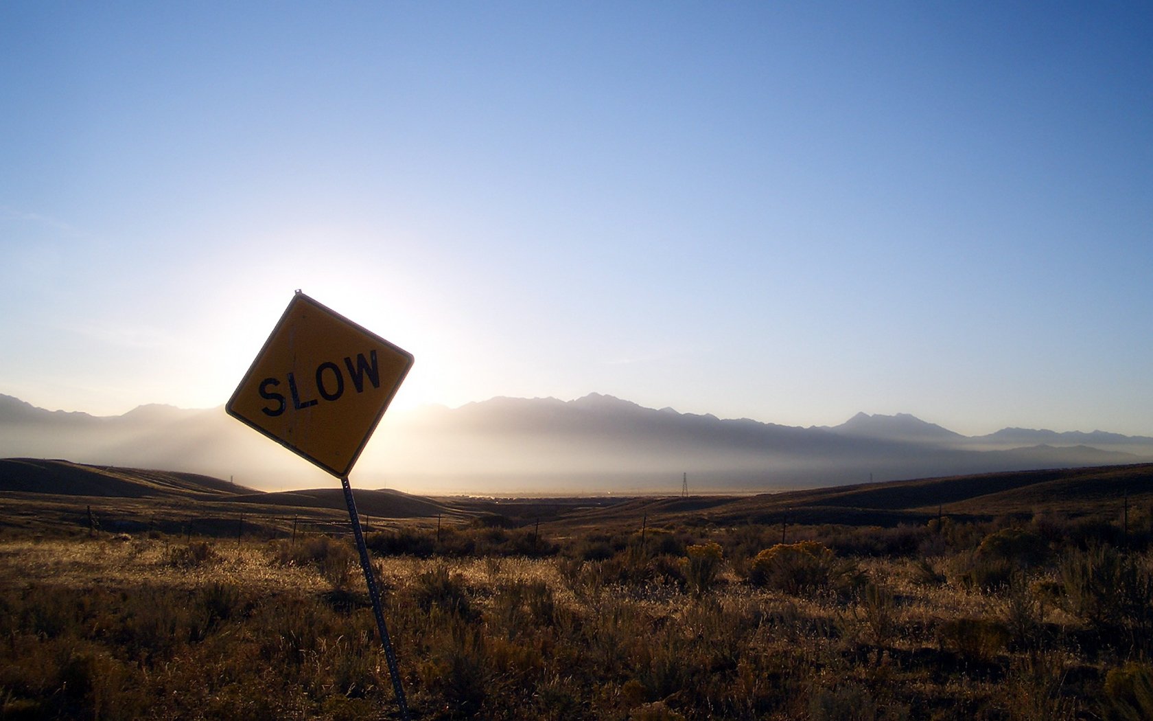 HD PC desktop wallpaper featuring a tilted SLOW road sign against a vast, sunlit desert landscape with distant mountains under a clear blue sky.