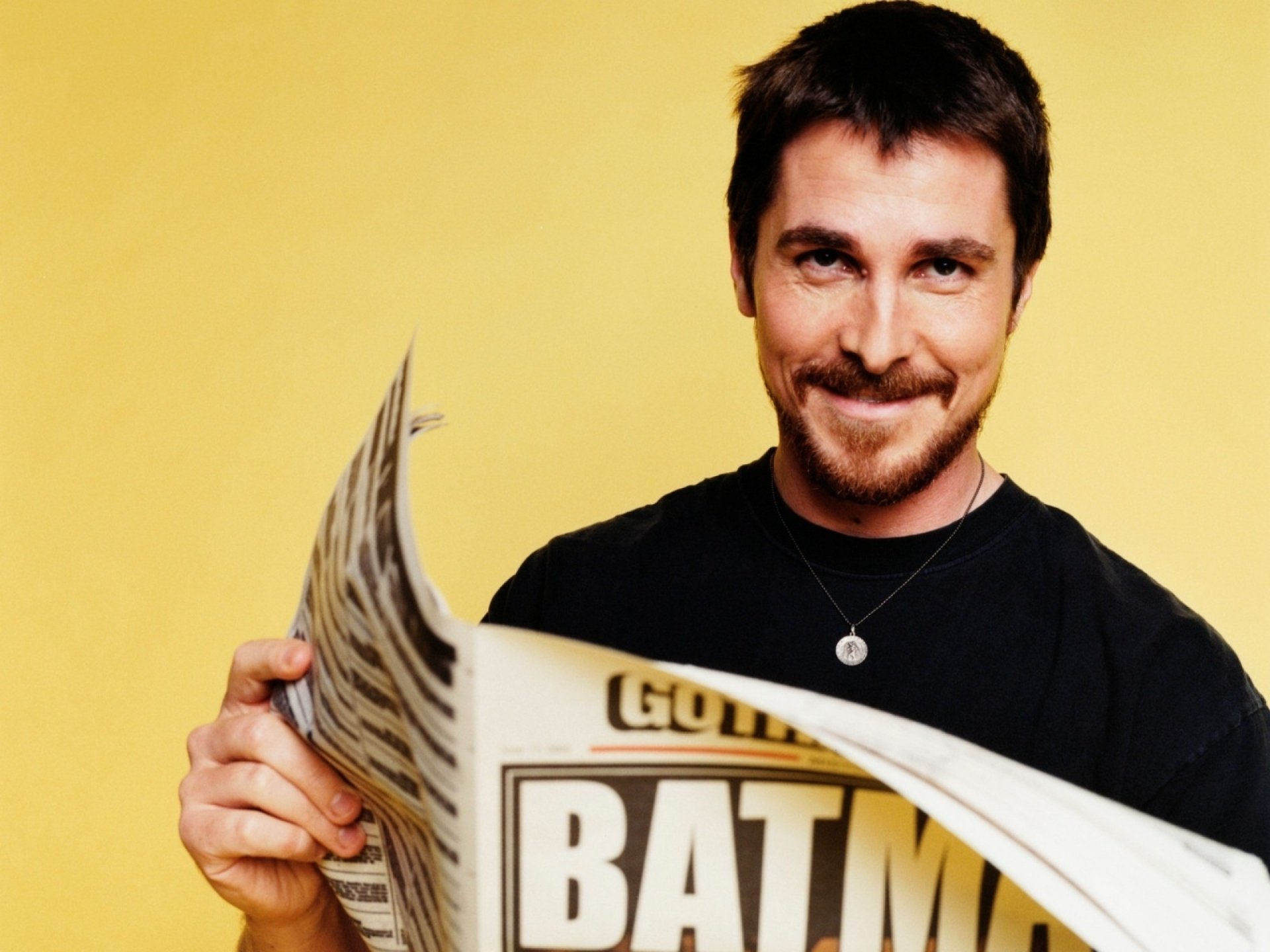 HD PC desktop wallpaper featuring celebrity Christian Bale smiling and holding a newspaper against a plain yellow background.