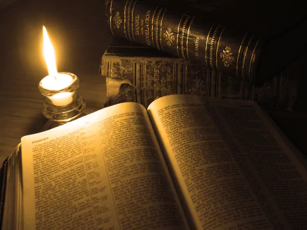 An open Bible rests on a table, illuminated by a flickering candle, surrounded by stacked religious books, creating a serene, contemplative atmosphere for this HD desktop wallpaper.