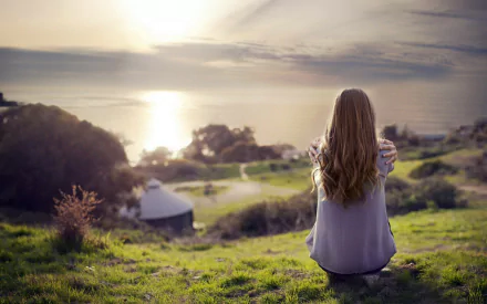 A woman with long hair, seen from the rear, sits on a hill overlooking a serene sunset by the sea. This HD desktop wallpaper and background exudes tranquility and beauty.