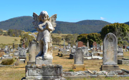 HD desktop wallpaper featuring a serene cemetery with a detailed angel statue standing among gravestones under a clear blue sky and distant hills.