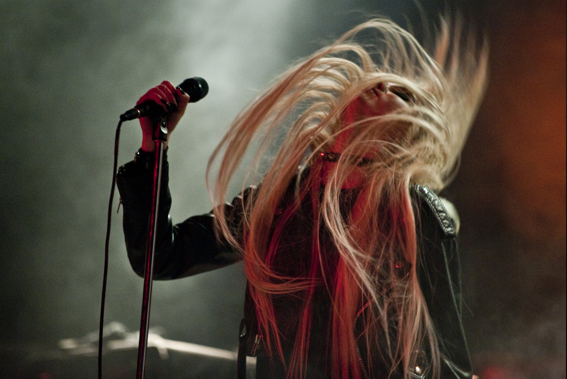 Concert music shot: blonde rock singer whipping long hair back while gripping a microphone amid smoky, moody stage lights — 4K Ultra HD PC desktop wallpaper.