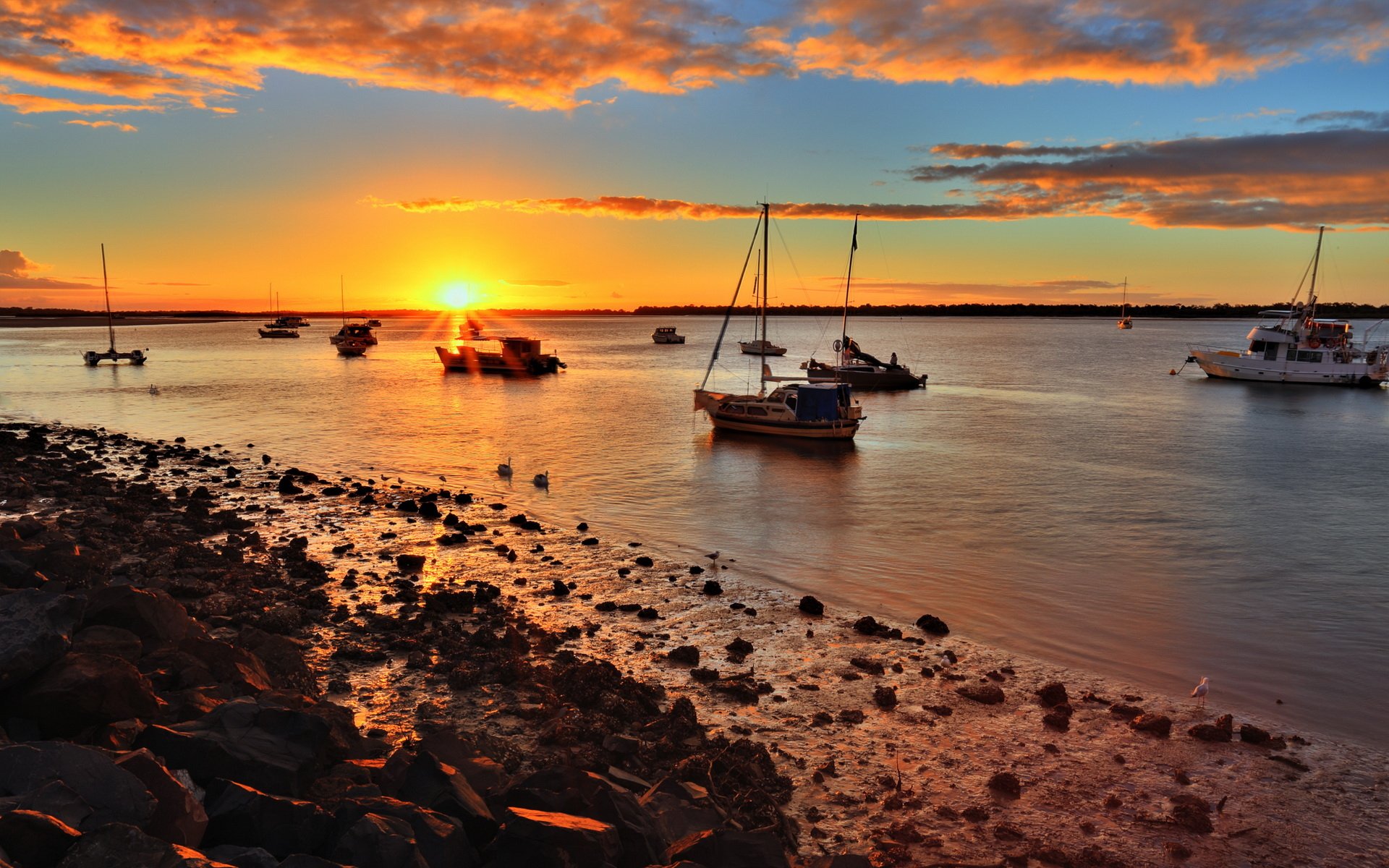 Photography sunset HD PC desktop wallpaper and background: golden sun sinking over a calm harbor of anchored sailboats and a rocky shoreline under colorful clouds.