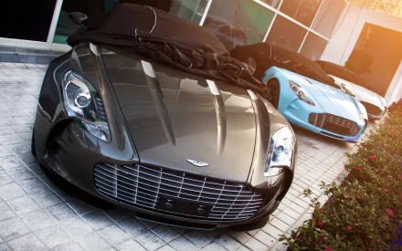 HD desktop wallpaper featuring two Aston Martin One-77 vehicles parked side by side, highlighted by polished surfaces and warm sunlight reflections.