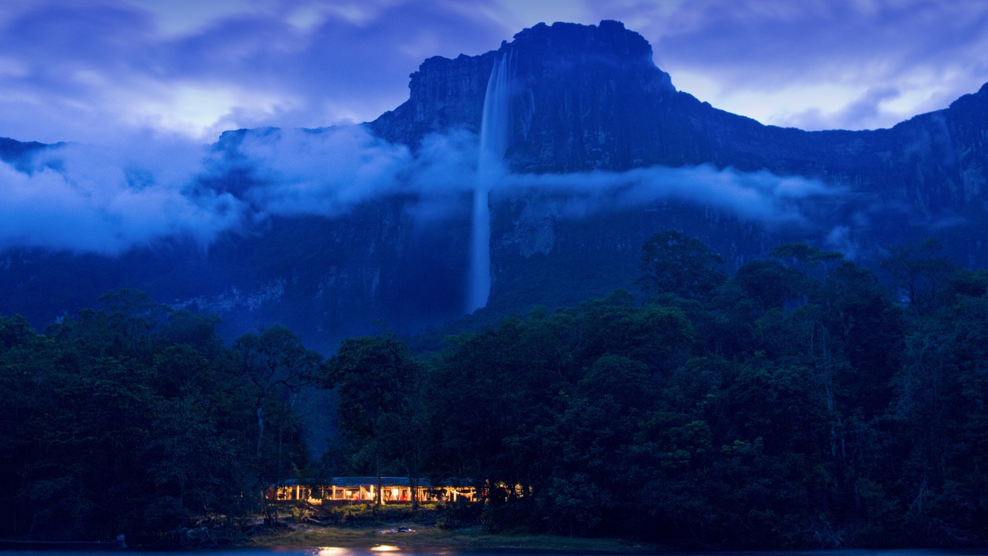 A serene view of Angel Falls cascading from a rugged mountain, surrounded by lush greenery and mist, featuring a warm glow from a nearby lodge under a twilight sky.