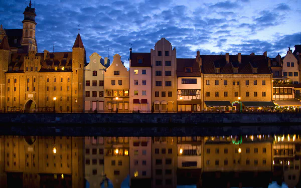 HD desktop wallpaper showcasing the illuminated, historic man-made waterfront buildings of Gdansk at dusk, reflected on calm water under a cloudy sky.