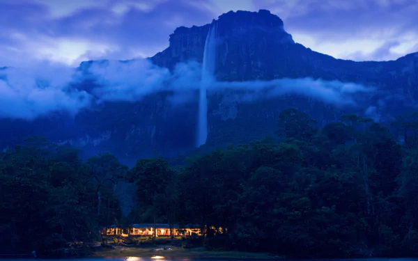 A serene view of Angel Falls cascading from a rugged mountain, surrounded by lush greenery and mist, featuring a warm glow from a nearby lodge under a twilight sky.
