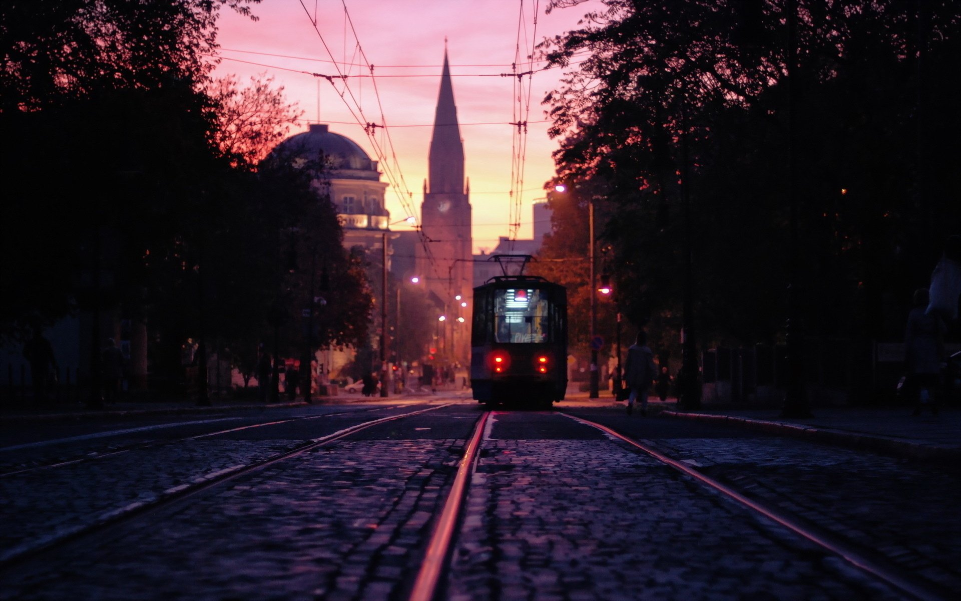 HD desktop wallpaper of a tram traveling on city tracks at sunset, with silhouetted buildings and a vibrant pink sky in the background.