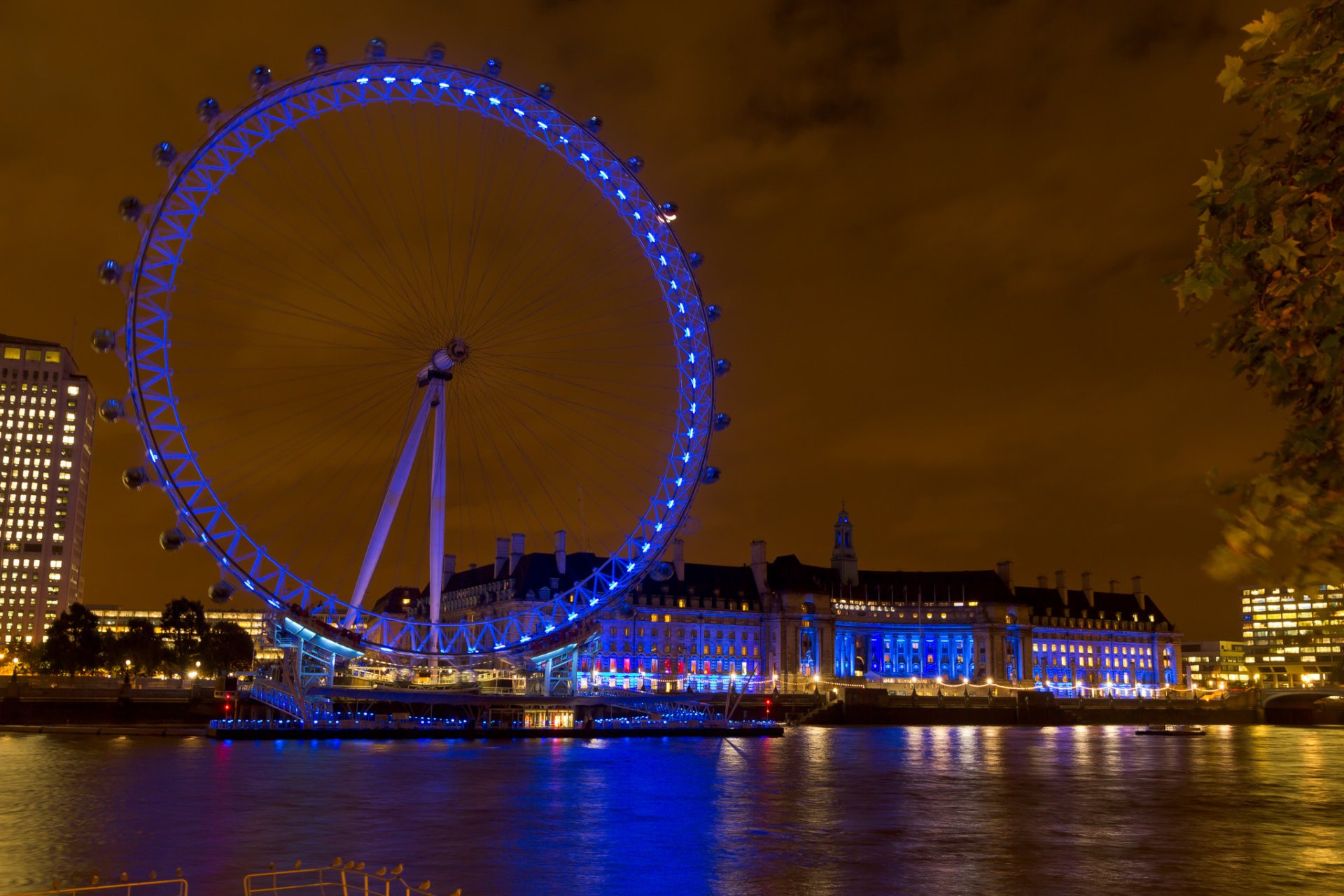HD PC desktop wallpaper showing a man-made Ferris wheel lit in blue at night, reflecting on the river with riverside buildings and an orange sky.