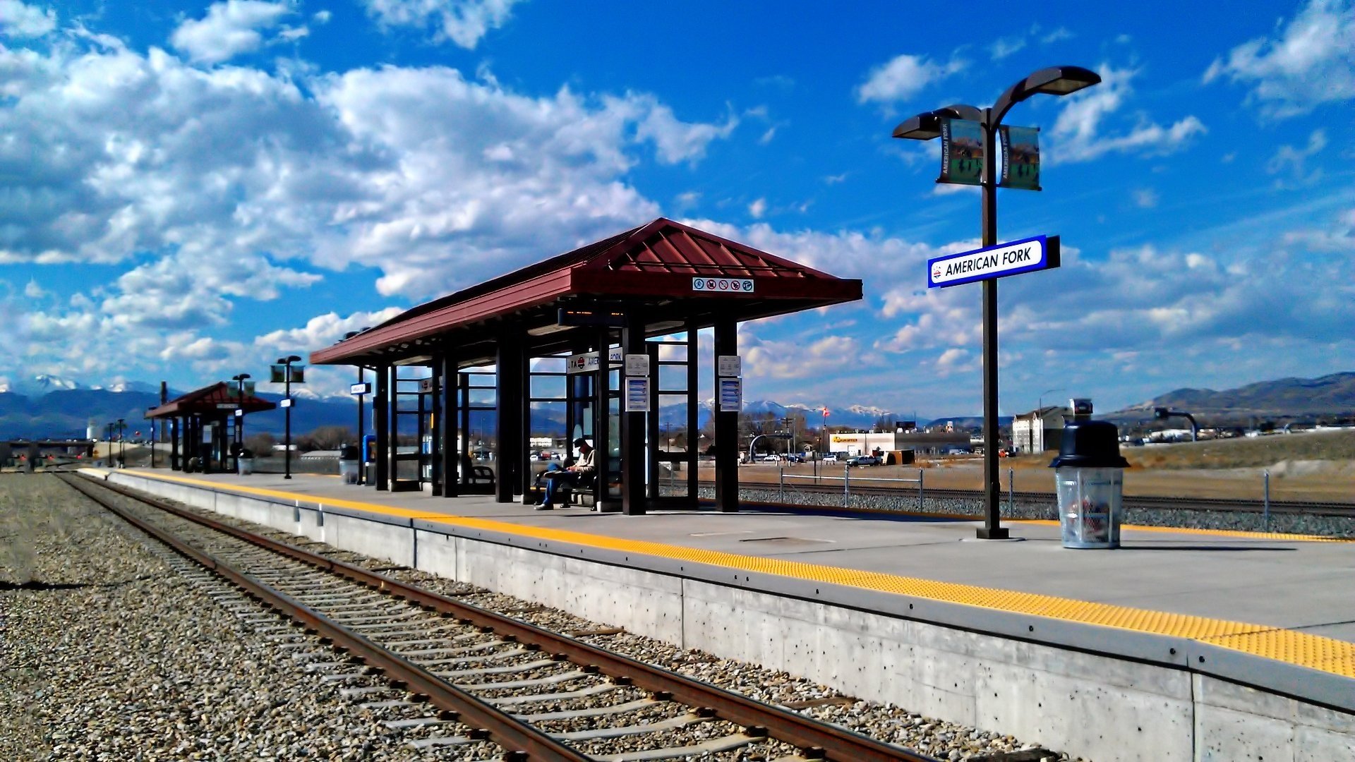HD desktop wallpaper featuring a serene train station with a platform under a clear sky and scattered clouds. The station is open with minimal structures, signage, and rail tracks in view.