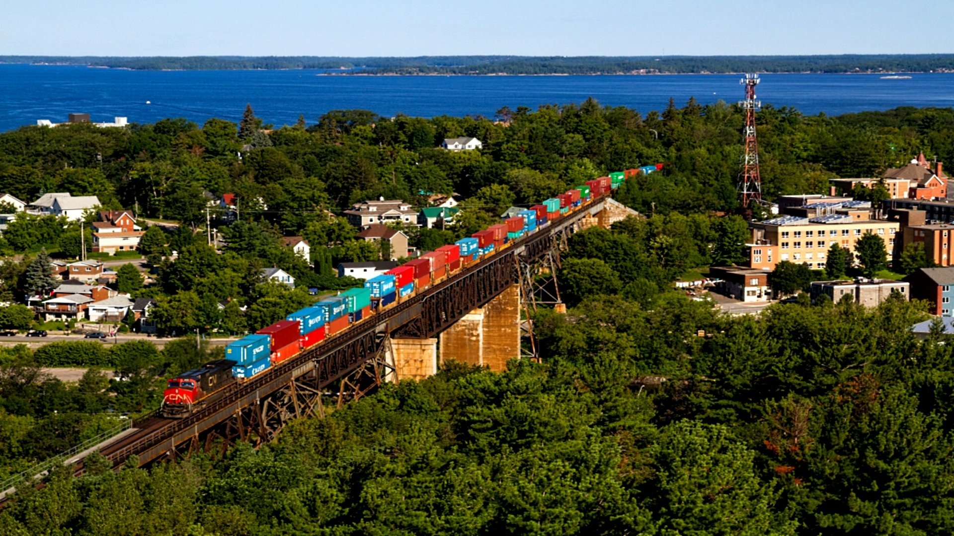 A train carrying colorful cargo containers crosses a bridge surrounded by lush greenery, with a serene body of water and urban landscape in the background. HD desktop wallpaper and background.