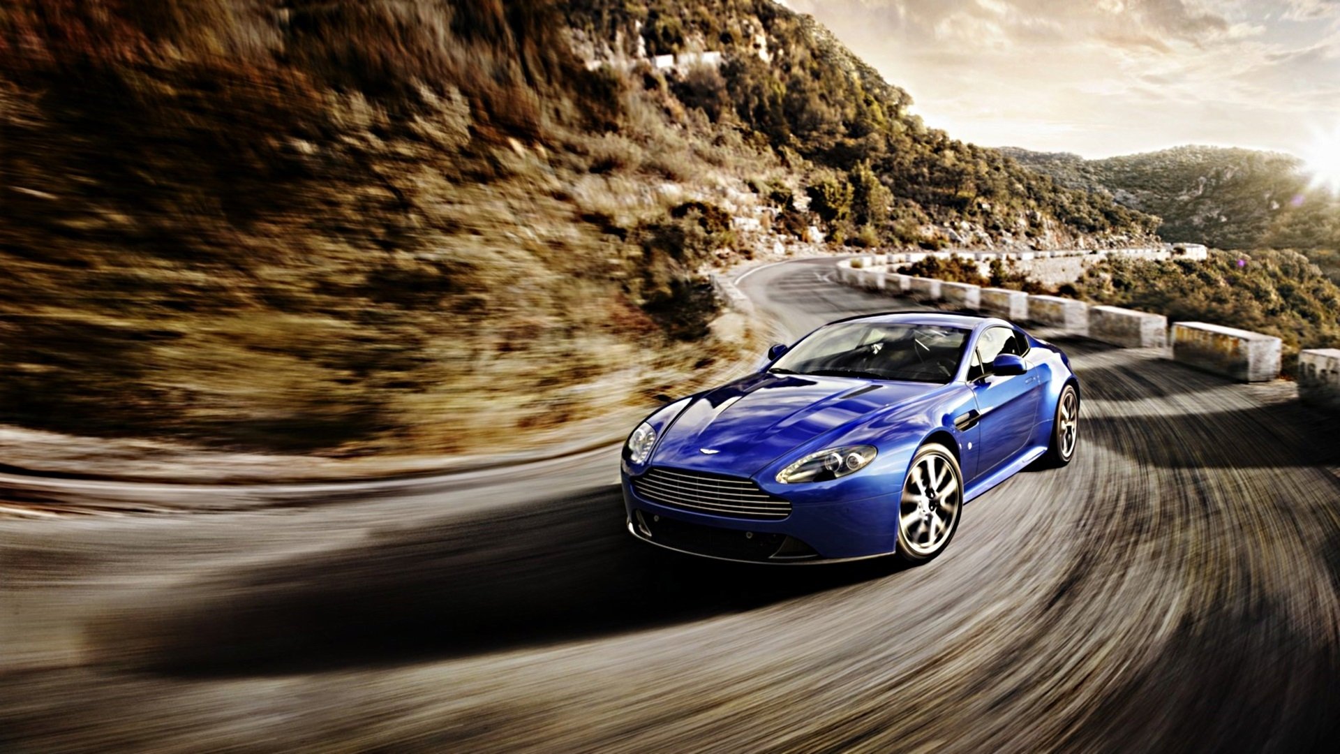 HD PC desktop wallpaper featuring a sleek blue Aston Martin V8 Vantage speeding along a winding mountain road surrounded by rocky cliffs and trees.