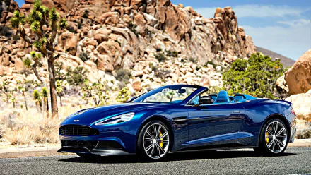 HD PC desktop wallpaper featuring a sleek blue Aston Martin Vanquish Volante convertible parked on a desert road with rocky hills and clear skies in the background.
