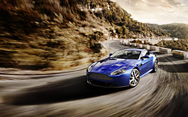 HD PC desktop wallpaper featuring a sleek blue Aston Martin V8 Vantage speeding along a winding mountain road surrounded by rocky cliffs and trees.