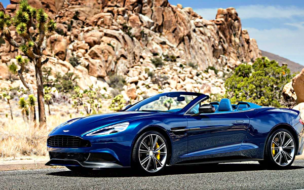 HD PC desktop wallpaper featuring a sleek blue Aston Martin Vanquish Volante convertible parked on a desert road with rocky hills and clear skies in the background.