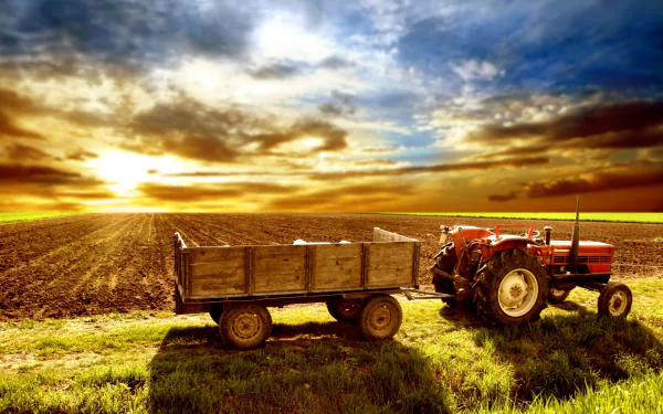 A red tractor with a wooden trailer stands in a vast field at sunset, under a dramatic sky. This HD desktop wallpaper captures the essence of rural farming life.