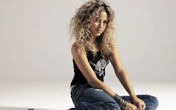 HD PC desktop wallpaper featuring Shakira with curly hair, sitting casually in a black top and jeans, representing music and style.
