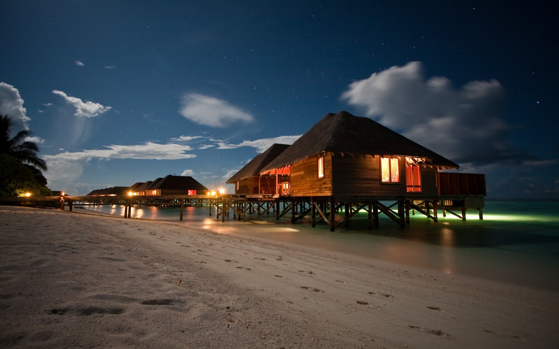 HD desktop wallpaper featuring a man-made cabin on stilts over calm ocean waters at night, with a sandy beach and illuminated rooms under a cloudy sky.