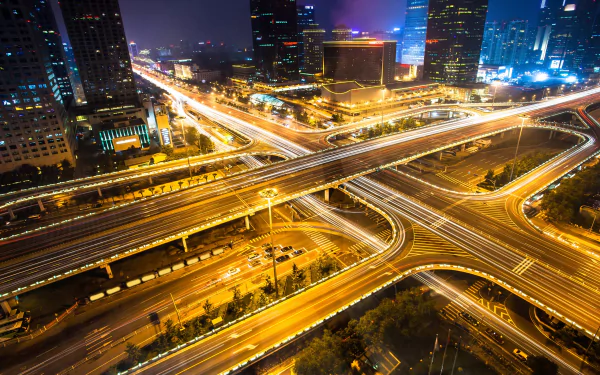 Photography time-lapse of a neon-lit urban highway interchange at night, long-exposure light trails weaving through the intersection — 2K Quad HD PC desktop wallpaper and background.