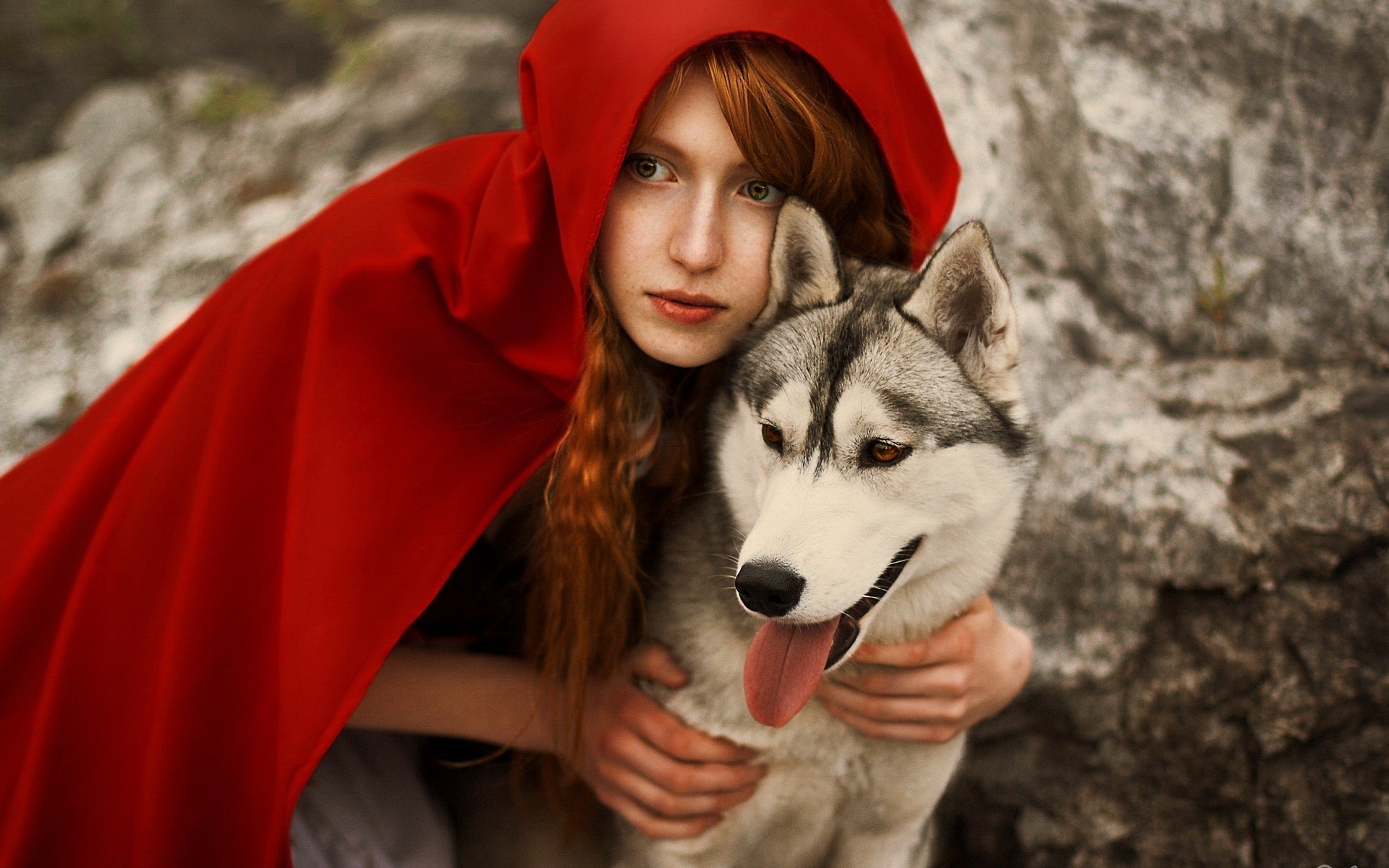 A woman in a vibrant red cloak poses with a husky dog against a rocky backdrop. This enchanting cosplay scene makes for a captivating HD desktop wallpaper and background.