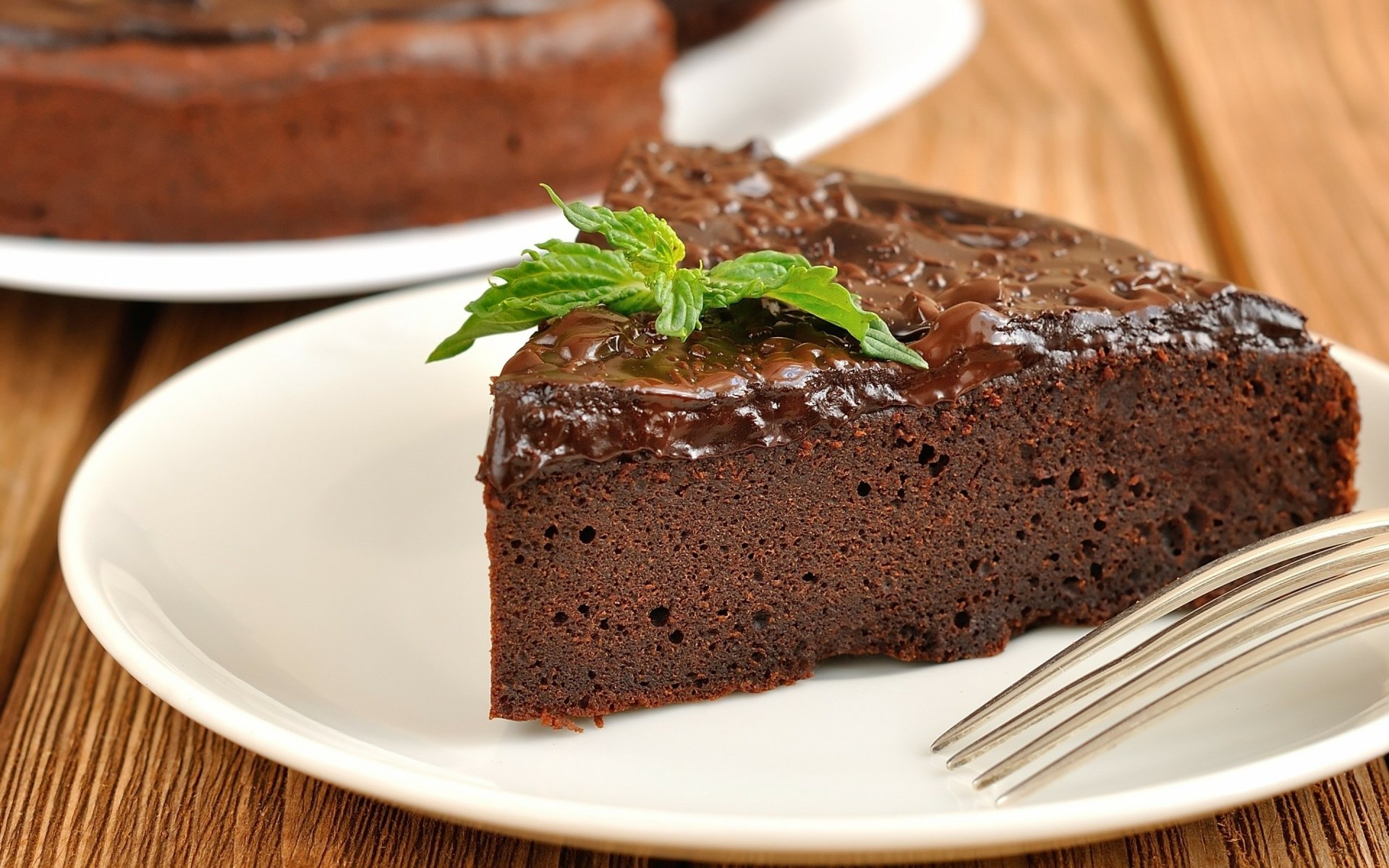 A delicious slice of chocolate cake garnished with mint on a white plate, set against a wooden background, creating an inviting food-themed HD wallpaper.