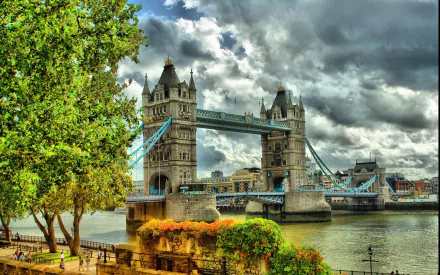 HD desktop wallpaper featuring London's iconic man-made Tower Bridge over the River Thames under a dramatic cloudy sky with lush greenery in the foreground.