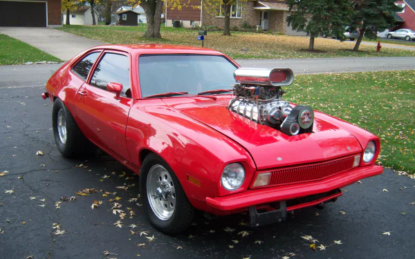 HD desktop wallpaper featuring a bright red Ford Pinto with a prominent supercharged engine parked on a driveway in a suburban neighborhood.